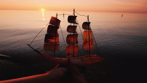 hands of a young couple raise up a model of a sailing ship on the background of the sea and sunset c