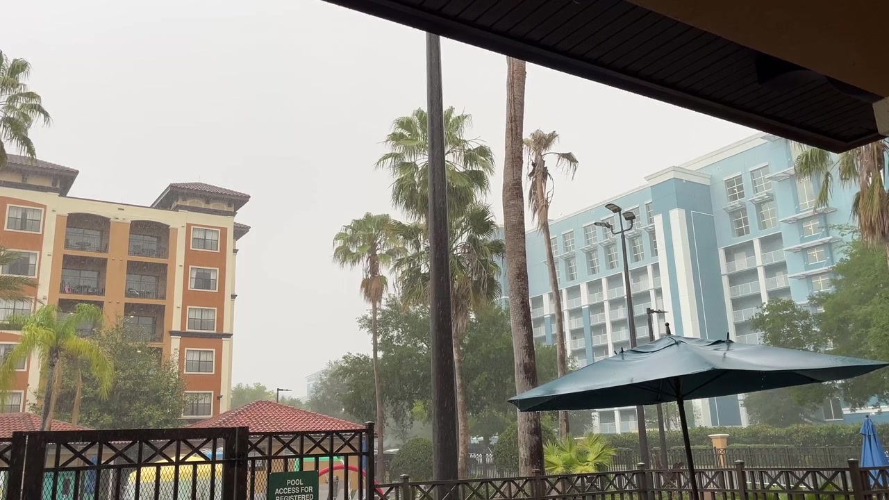 Tormenta de lluvia cayendo sobre palmeras y edificios de resort junto a una zona de piscina vallada. Amplia vista exterior mostrando lluvia intensa y clima tropical en un complejo hotelero