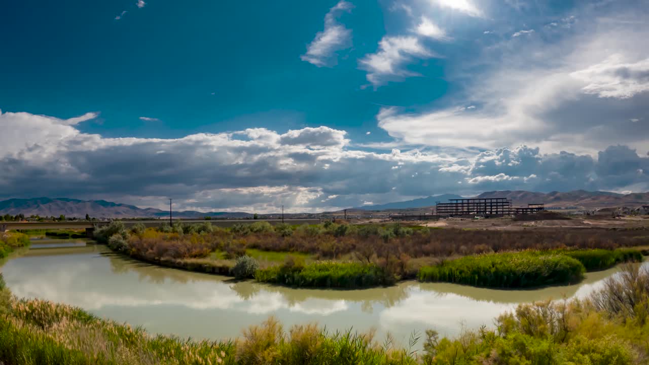 paisaje nublado sobre un río con el reflejo del cielo en el agua y una carretera y un proyecto de construcción junto a las montañas distantes - alejar el lapso de tiempo