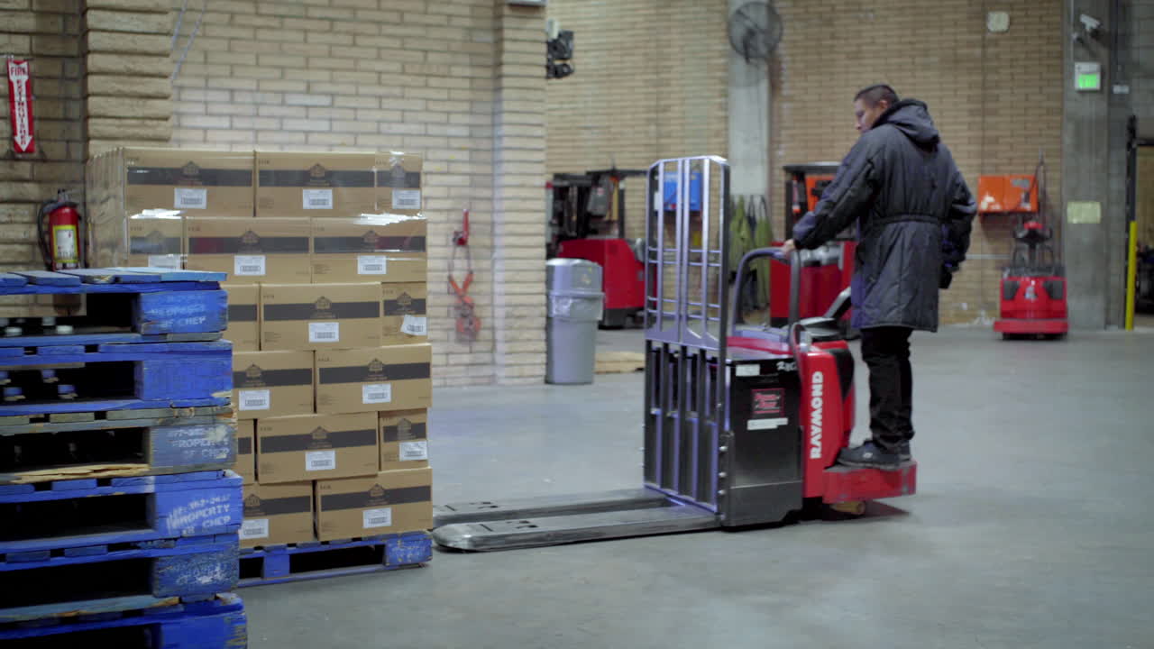 A worker operates a pallet jack, moving a stack of boxes in a warehouse