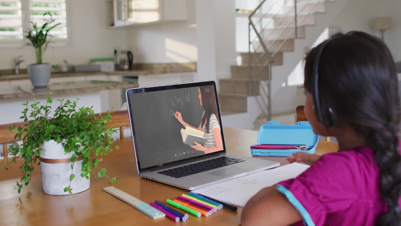 Niña afroamericana haciendo la tarea mientras tiene una videollamada con una profesora en el portátil en casa.