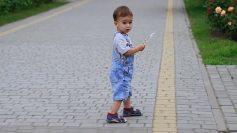 adorable little kid in a jeans romper walking by the road. kid holding a magazine in his hands comes up to a big statue of a cat carved from wood.