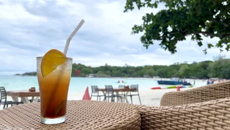 a cold cocktail with an orange slice and straw sits on a wicker table overlooking a sandy beach and calm sea under cloudy daylight