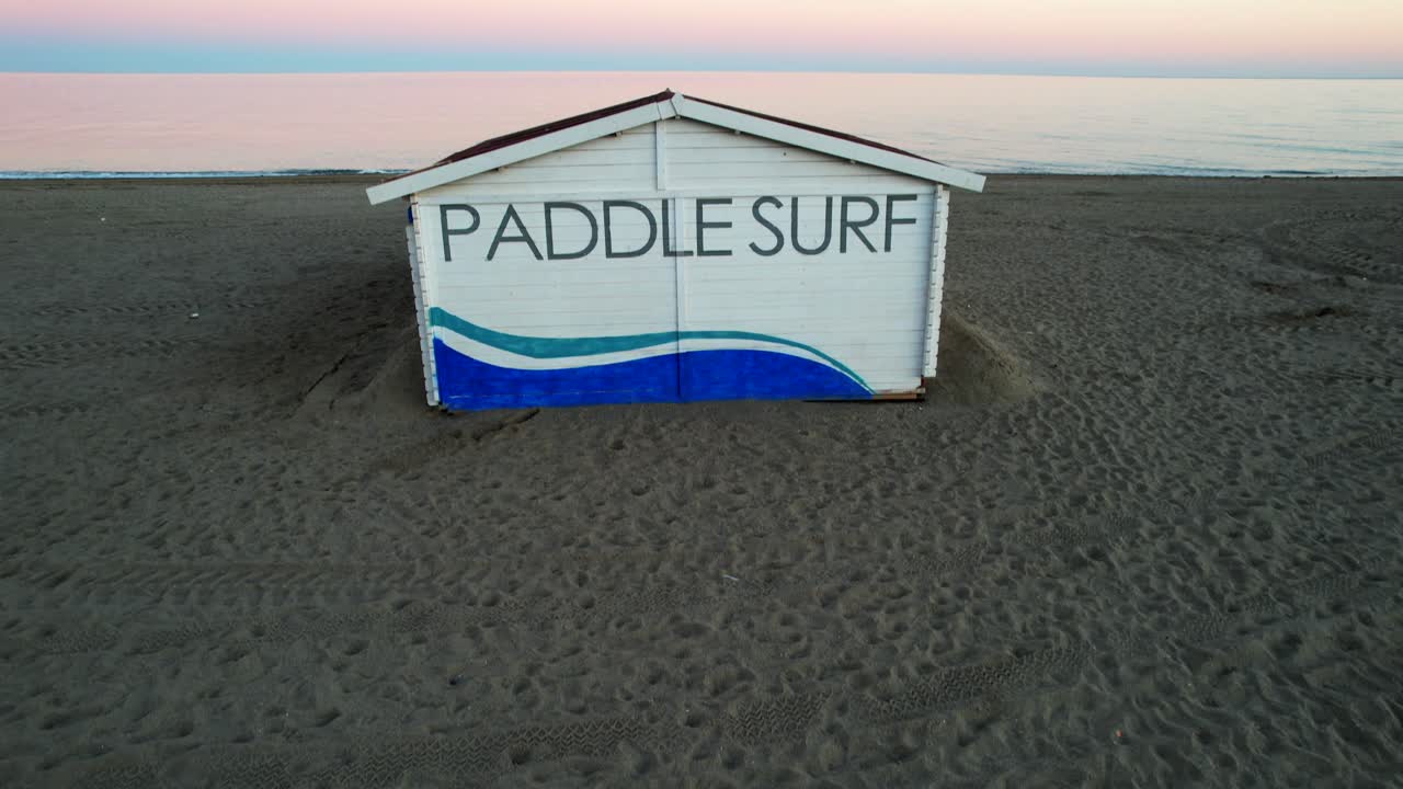 Aerial tilt up of shack with paddle surf words, pastel sunset sky behind
