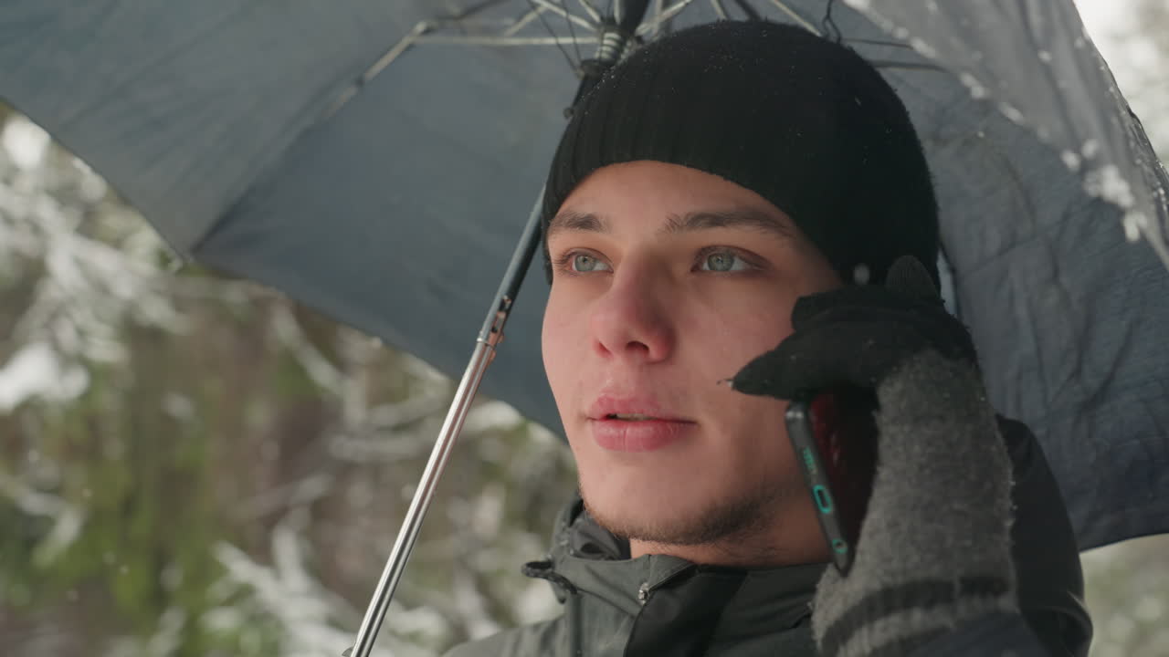 Young white man under umbrella calling, standing in snowy forest, wearing black beanie and puffer jacket, gray gloves holding smartphone to ear, breath visible, snowflakes falling, introspective