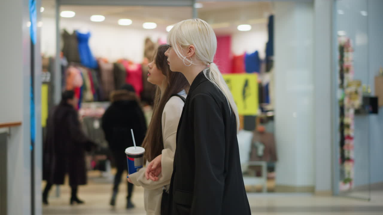 Side view of ladies walking toward ascending escalator in modern shopping mall, one holding drink cup, stylish outfits and background with shoppers and clothing stores