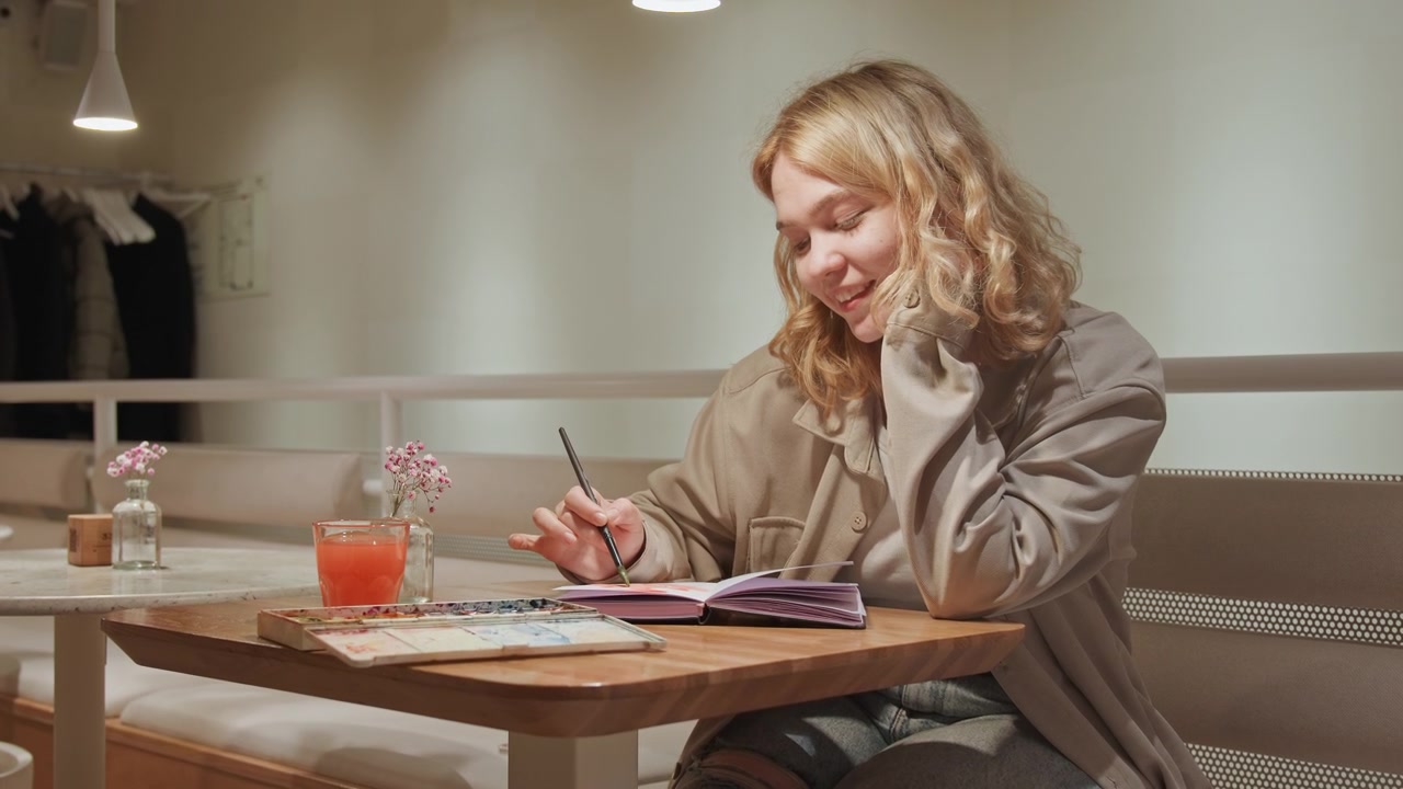 Primo piano di una giovane donna che dipinge in un caffè d'arte, tiene un pennello e un album da disegno con una tavolozza di acquerelli sul tavolo. Ambiente interno luminoso con mobili in legno, un piccolo vaso di vetro e fiori rosa.