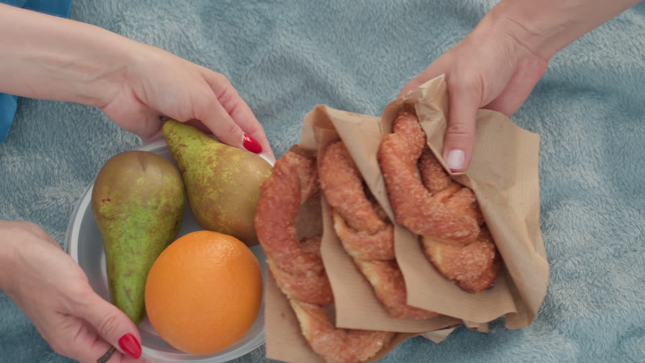 high angle view of mother hand placing baked bread and plate of orange on picnic mat, soft blanket on grass, summer park setting, preparing snack, warm light, casual outdoor gathering