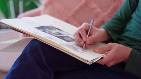 close up of arstist's hands drawing woman's face on sketch pad while sitting on pink couch