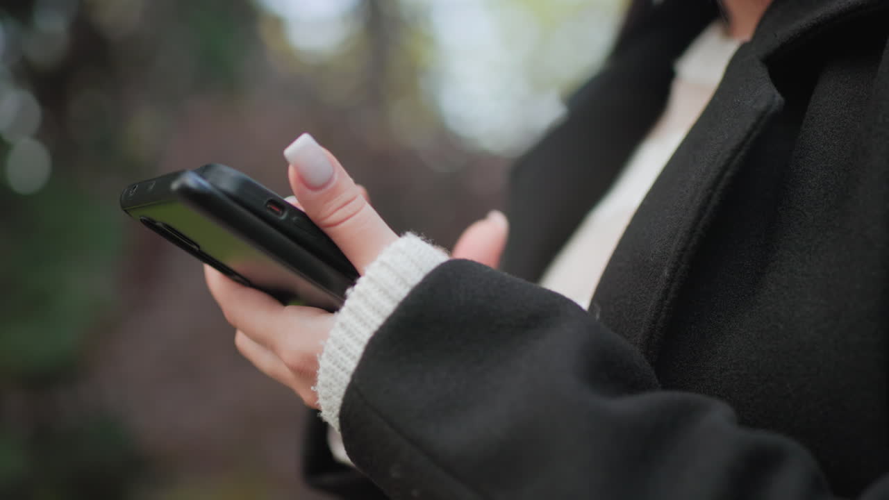 야외에서 휴대폰을 들고 있는 손 관리 네일, 부드러운 보케 배경, 아늑한 코트 소매, 부드러운 터치 상호 작용, 공원 일광 아래 현대적인 소통 순간