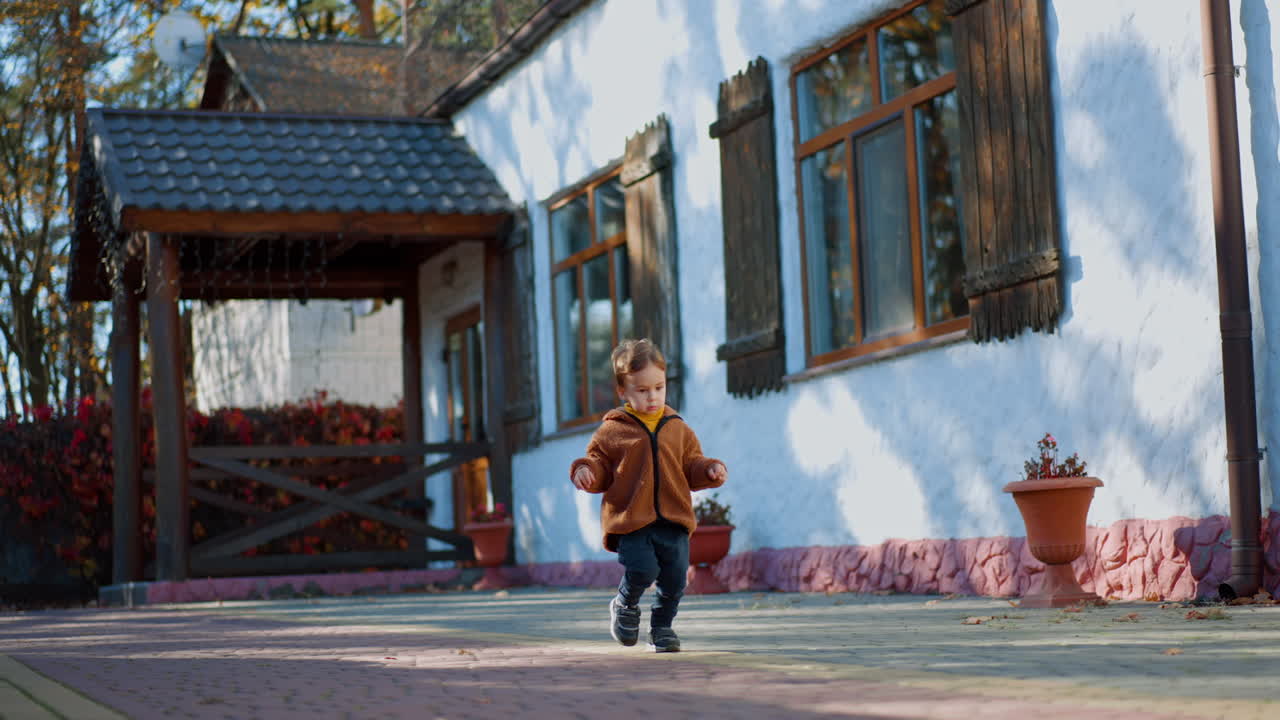 Caucasian toddler wearing brown jacket and blue pants runs outdoors. Good time outside in autumn.