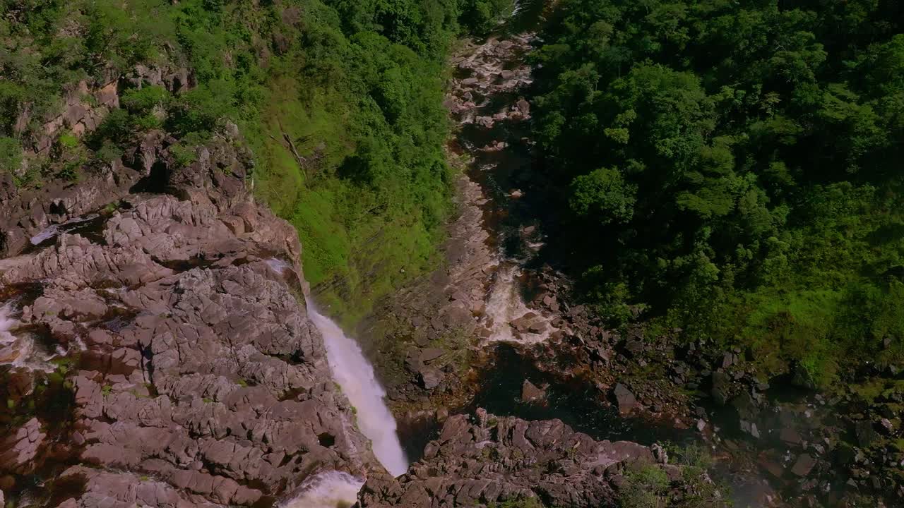 Stunning aerial view of a waterfall cascading down rocky cliffs in a lush green forest in Chapada Dos Veadeiros, Brazil, creating a rainbow in the mist, slow moton drone shot