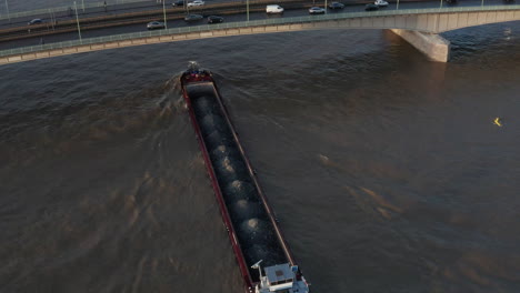 High-angle-view-of-loaded-cargo-ship-passing-under-busy-road-bridge-at-golden-hour.-Cologne,-Germany