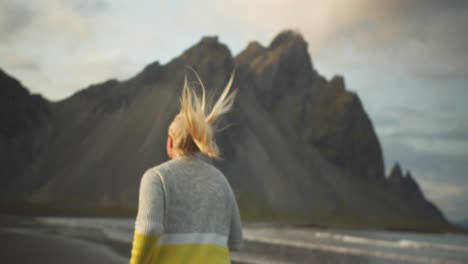 young blonde woman runs athletically along the shore and looking back laughing in front of rocky mountain landscape