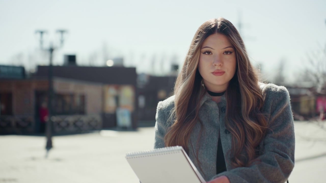 Donna urbana che esamina i modelli di abbigliamento. Scena all'aperto di donna che analizza illustrazioni di moda. Sfondo di paesaggio urbano con donna che esamina schizzi all'esterno, indossando un cappotto invernale e con i capelli lunghi.