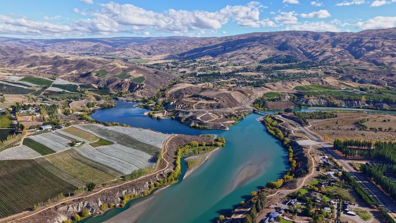 Drone ascends and pulls back above turquoise river, bridge, and farmlands under blue sky