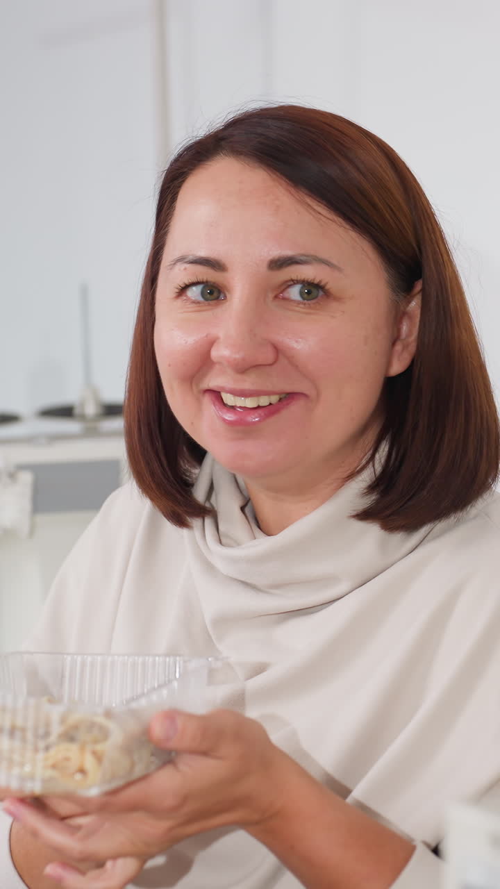 Una mujer comiendo un pastel en casa, sonriendo mientras sostiene un recipiente de plástico transparente en una cocina acogedora, pausa relajada por la tarde, retrato sincero y amigable con un suéter informal, saboreando un dulce con satisfacción.