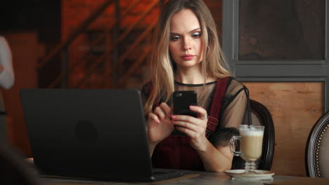 young woman sitting in coffee shop at wooden table, drinking coffee and using smartphone.on table is laptop. girl browsing internet, chatting, blogging. female holding phone and looking on his screen.