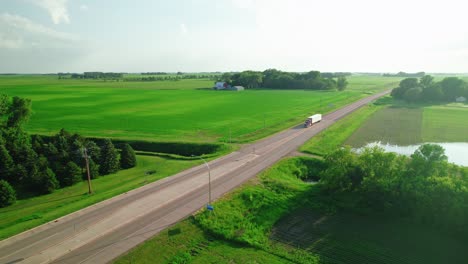 static aerial in the morning view of a red semi-truck driving on a rural highway through green farmland.