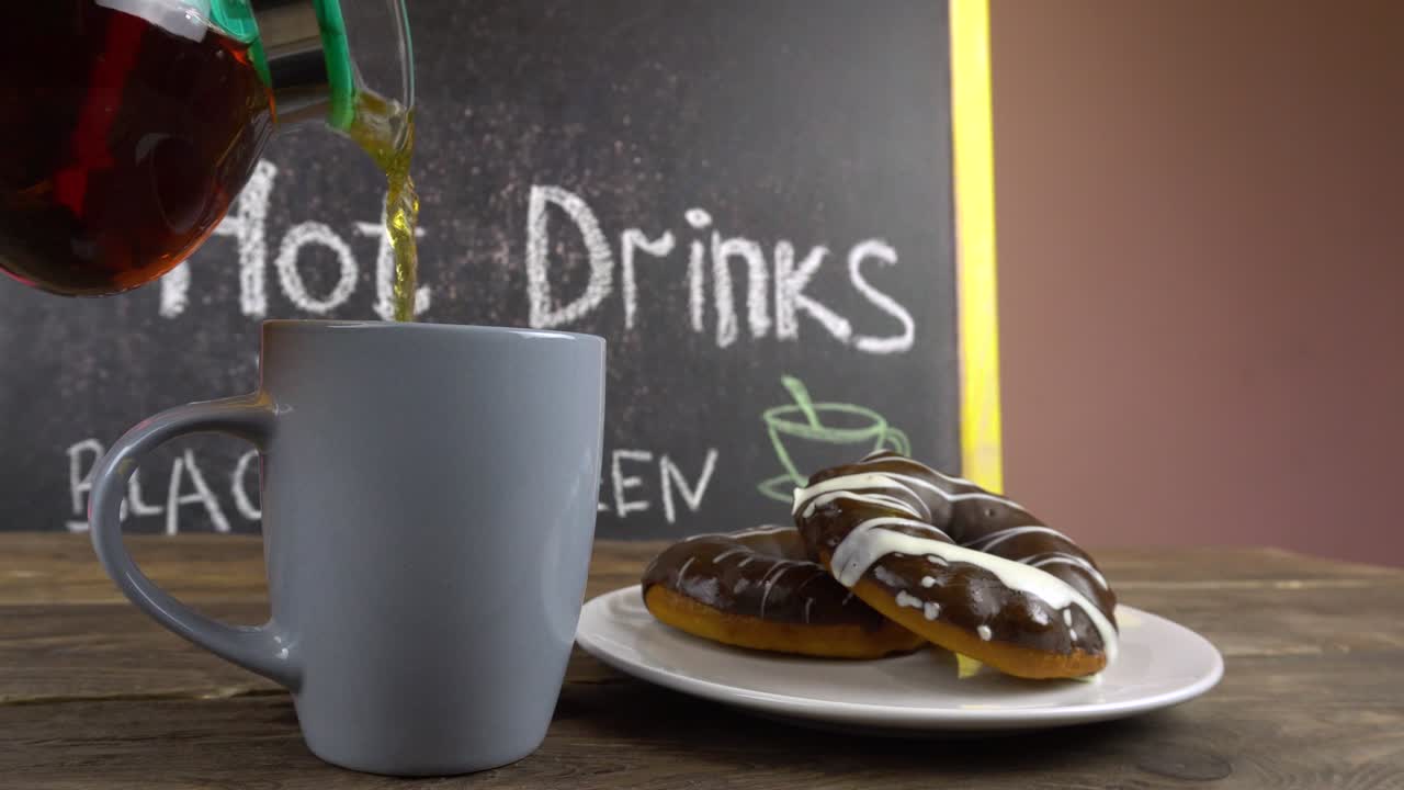 una taza de té y rosquillas en glaseado de chocolate en una mesa de café. el té está derramando de una tetera transparente.