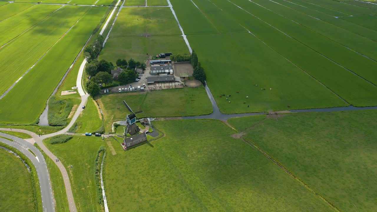 Scenic drone footage showing a traditional Dutch windmill located at a rural intersection, surrounded by expansive green farmland and cycling paths