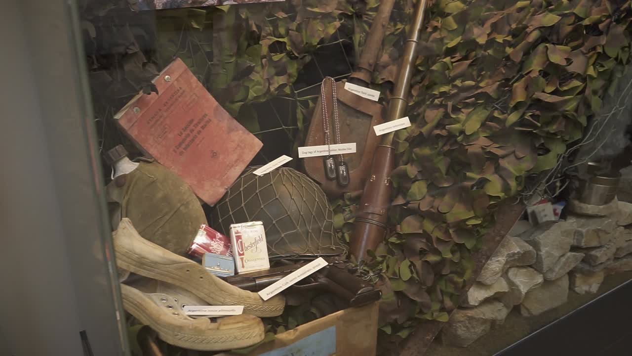 Military artifacts from world war ii, including a helmet, canteen, rifle, documents, cigarettes, and camouflage netting, are showcased in a museum display case