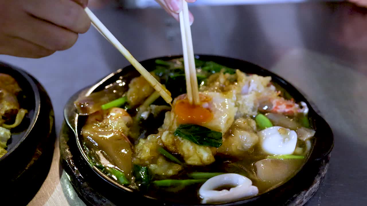 Chopsticks lift seafood pad thai noodles from a sizzling hot plate under bright lighting
