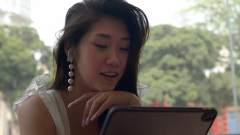 portrait of beautiful young charming asian girl having internet call on her tablet, smiling in coffee shop