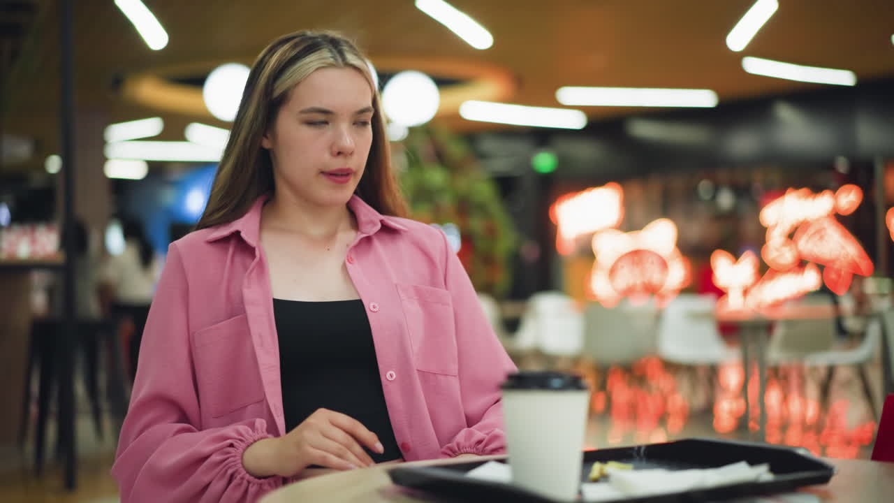 mujer de vestido rosa disfrutando del café, colocando la taza en una bandeja, luego relajándose con una expresión satisfecha, frotándose el estómago después de la comida, personas borrosas caminan en el fondo con luces de neón