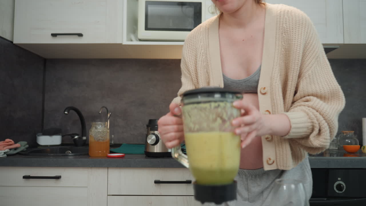 Mujer embarazada preparando un batido matutino en la cocina, siguiendo una receta en el teléfono, midiendo verduras y frutas, vistiendo un cárdigan con el vientre de embarazada visible, luz natural suave, ambiente doméstico relajado