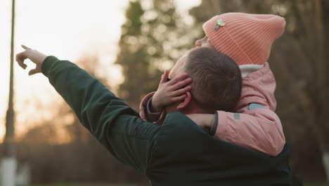 a man in a green jacket carries his daughter, who is dressed in a pink jacket and matching beanie, on his shoulders. the girl places her hand gently on his face as he points towards the sky