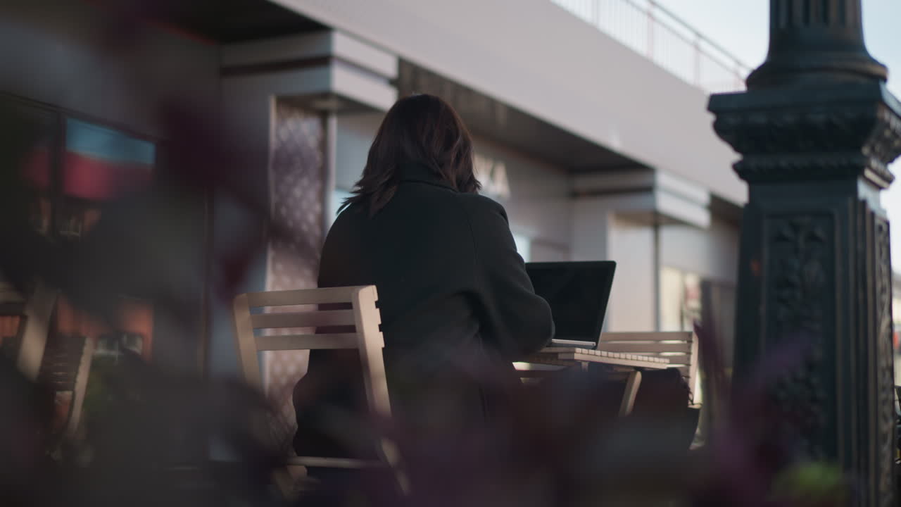 Lady in black outfit typing on laptop while seated outdoors at cafe table surrounded by wooden chairs and blurred plants in foreground