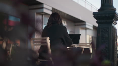 lady in black outfit typing on laptop while seated outdoors at cafe table surrounded by wooden chairs and blurred plants in foreground