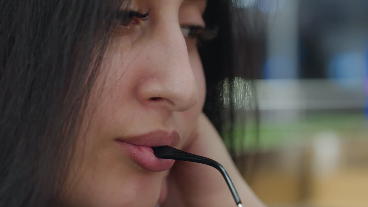 Close up of fair skin lady with long dark hair gently holding eyeglass frame in mouth while resting chin on hand, expressing soft charm and quiet thoughtfulness in cozy indoor environment