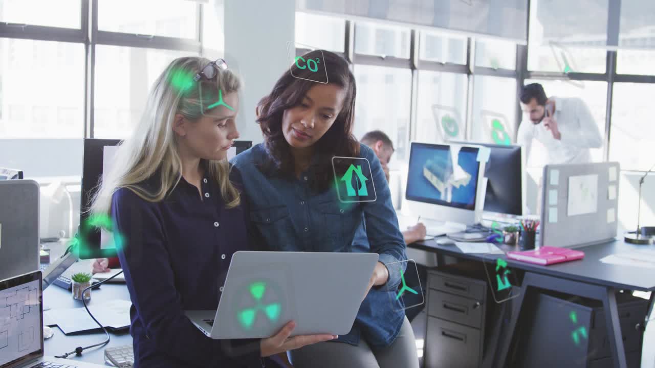 Equipo de diseño revisando una laptop, una persona de cabello oscuro señalando y tocando, mientras iconos verdes aparecen sobre los escritorios.