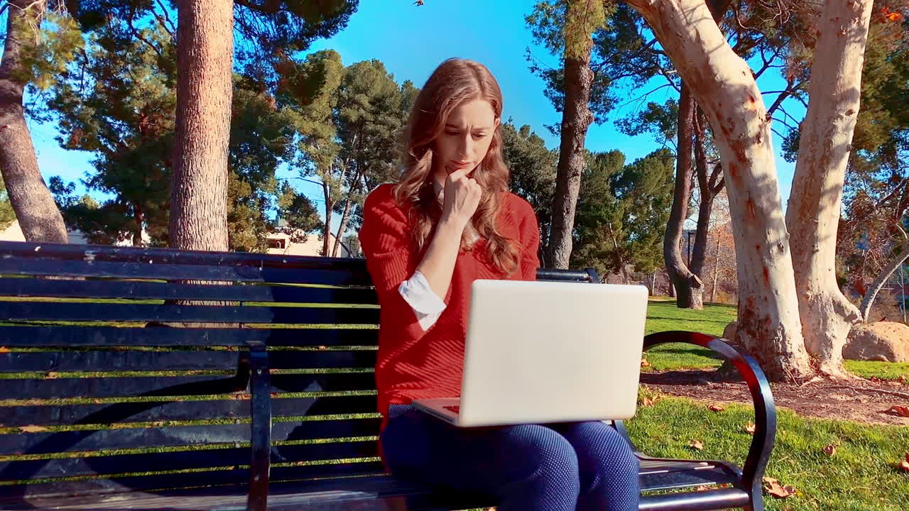 A happy, attractive, young, caucasian woman at the park, on a bench using her laptop