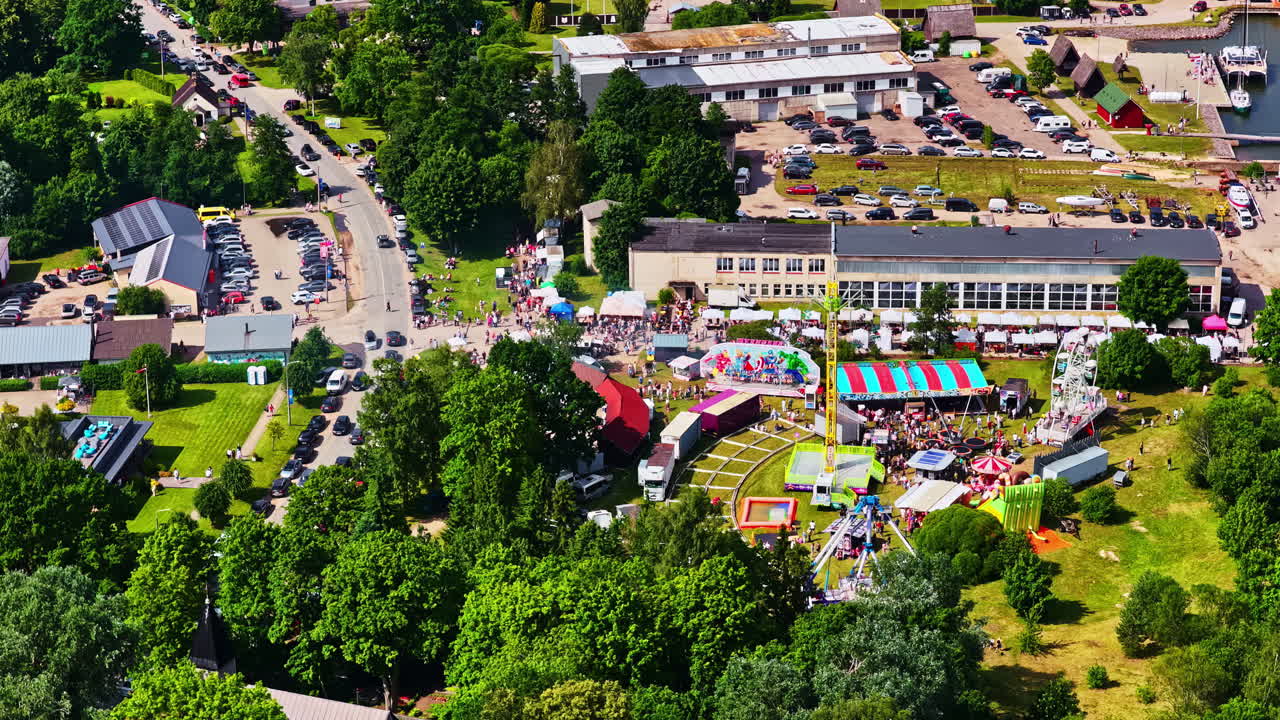 Aerial drone view of bustling Zvejnieksvētki, a traditional Fisherman's Festival, with carnival rides, tents, and crowds of people celebrating in Engure, Latvia