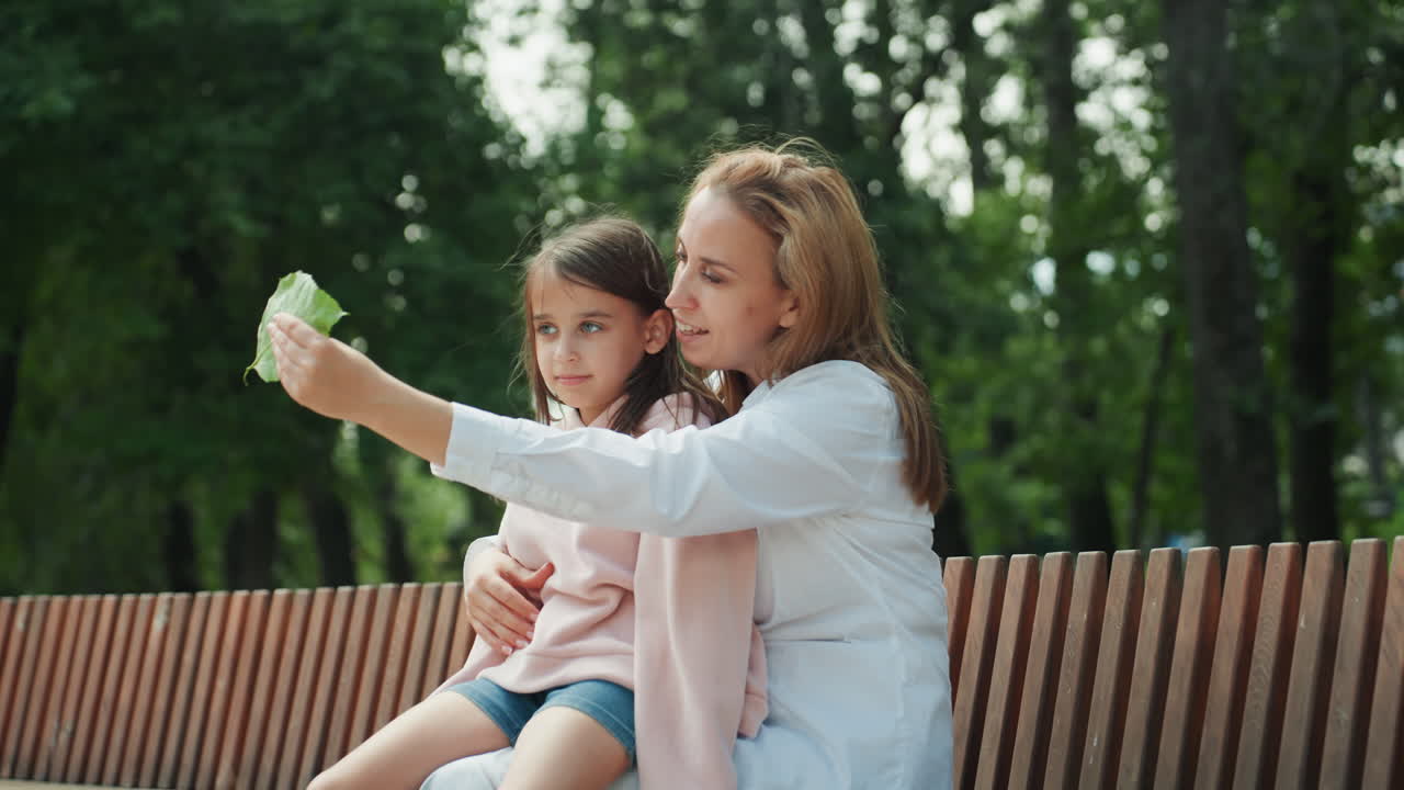 Mamma e figlia su una panchina del parco si protendono verso una foglia, in una scena di scoperta giocosa con la madre che incoraggia la curiosità. Lo sfondo è composto da alberi frondosi, con una luce naturale intensa, creando un momento interattivo di meraviglia e attenzione condivisa.