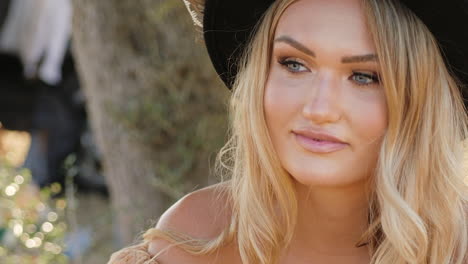 Beautiful-blonde-woman-resting-and-smiling-under-a-tree-in-a-boho-style-dress-and-fedora-hat---close-up