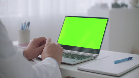 portátil con pantalla verde para tecnología de llave croma en la mesa del médico consultando en línea y viendo el seminario web el médico está trabajando de forma remota