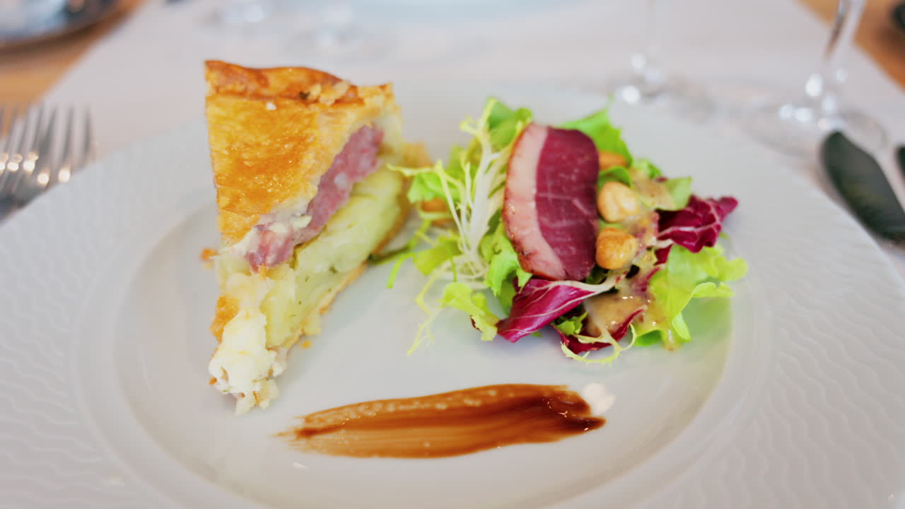 Close up of a slice of quiche featuring with a side salad and a slice of cured meat on a white plate at a restaurant