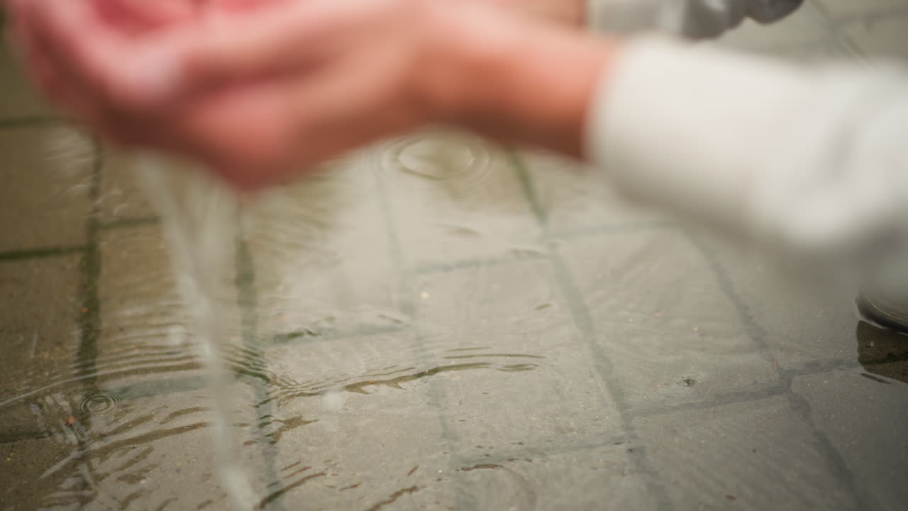 비 온 뒤 젖은 보도 타일을 손으로 닦는 모습, 물웅덩이의 잔물결과 흐린 반영이 있는 반사되는 콘크리트 위로 손바닥이 미끄러지는 클로즈업, 도시 환경에서 사색적인 배경 장면