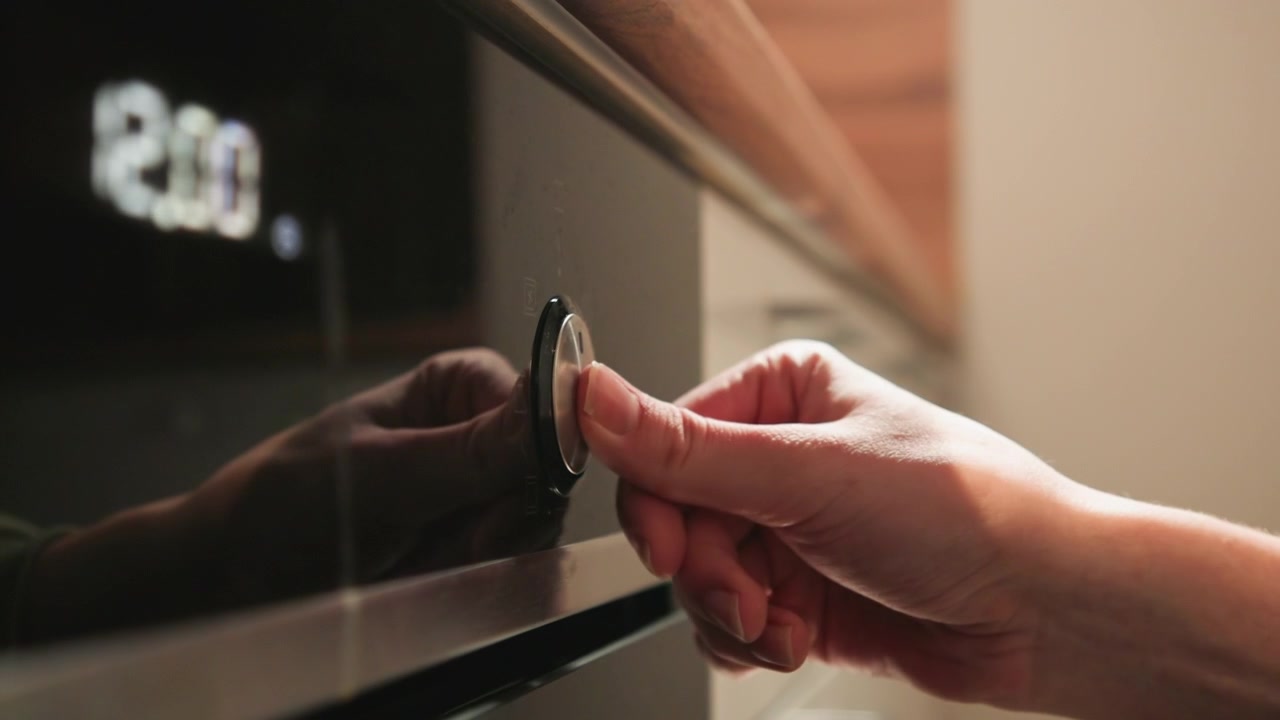 Primo piano dal basso di una mano che afferra e ruota una manopola di controllo cilindrica del forno accanto al display illuminato "230", con tenui riflessi sul pannello nero in acciaio inossidabile e una calda luce da cucina.
