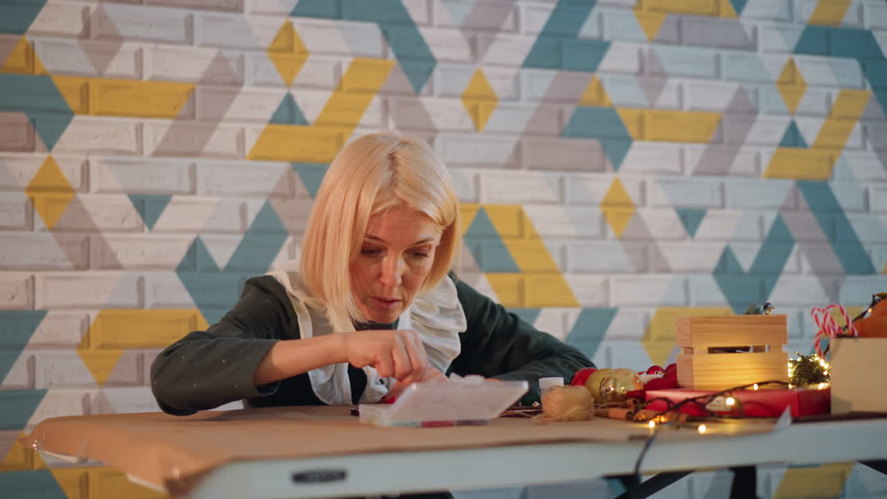 Donna caucasica che incarta un regalo di Natale su un moderno tavolo da studio, concentrata nell'annodare un nastro e scrivere un biglietto accanto a una piccola scatola dorata e lucine. Lo sfondo è una carta da parati geometrica, per un'atmosfera calda e accogliente.