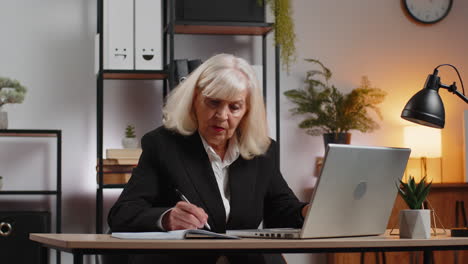 focused senior businesswoman writing notes and ideas in diary and using laptop at home office desk