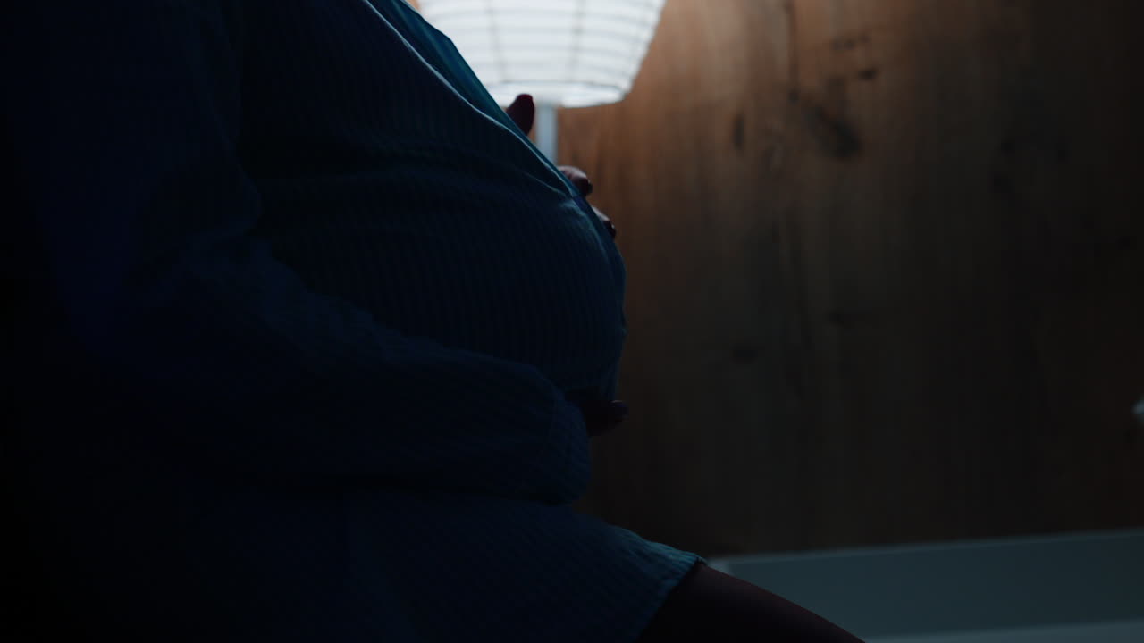 Pregnant belly of an unrecognized woman. Lady strokes her belly tenderly with love. Night lamp is on at backdrop.