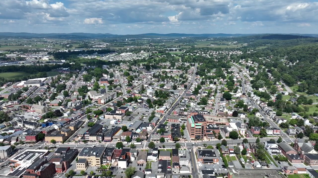 Aerial wide shot of cityscape in USA surrounded by green forest on cloudy day. Main street with traffic scene in Pennsylvania. Houses and buildings in city center.