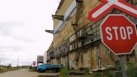 Vista-Exterior-De-La-Fábrica-Soviética-Abandonada-De-Fundición-De-Metalurgia-Pesada-Liepajas-Metalurgs-Territorio,-Señal-De-Alto-En-Los-Ferrocarriles,-Camión-De-Carga-Azul-En-El-Fondo,-Día-Nublado,-Plano-General