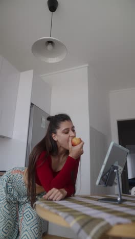 woman eating an apple while using a tablet in a kitchen