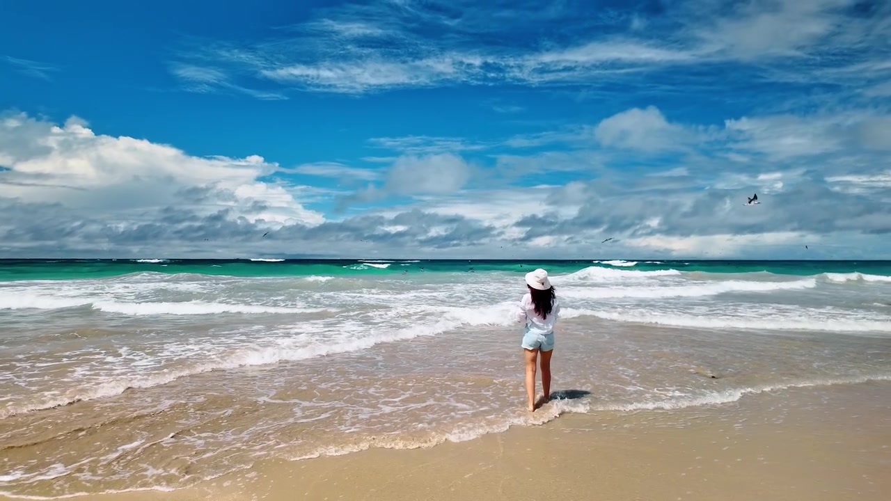 Mulher em pé na beira do mar na praia da Ilha Isabela. Vista traseira de uma viajante segurando seu chapéu enquanto ondas espumosas cruzam a areia bronzeada sob faixas de nuvens.